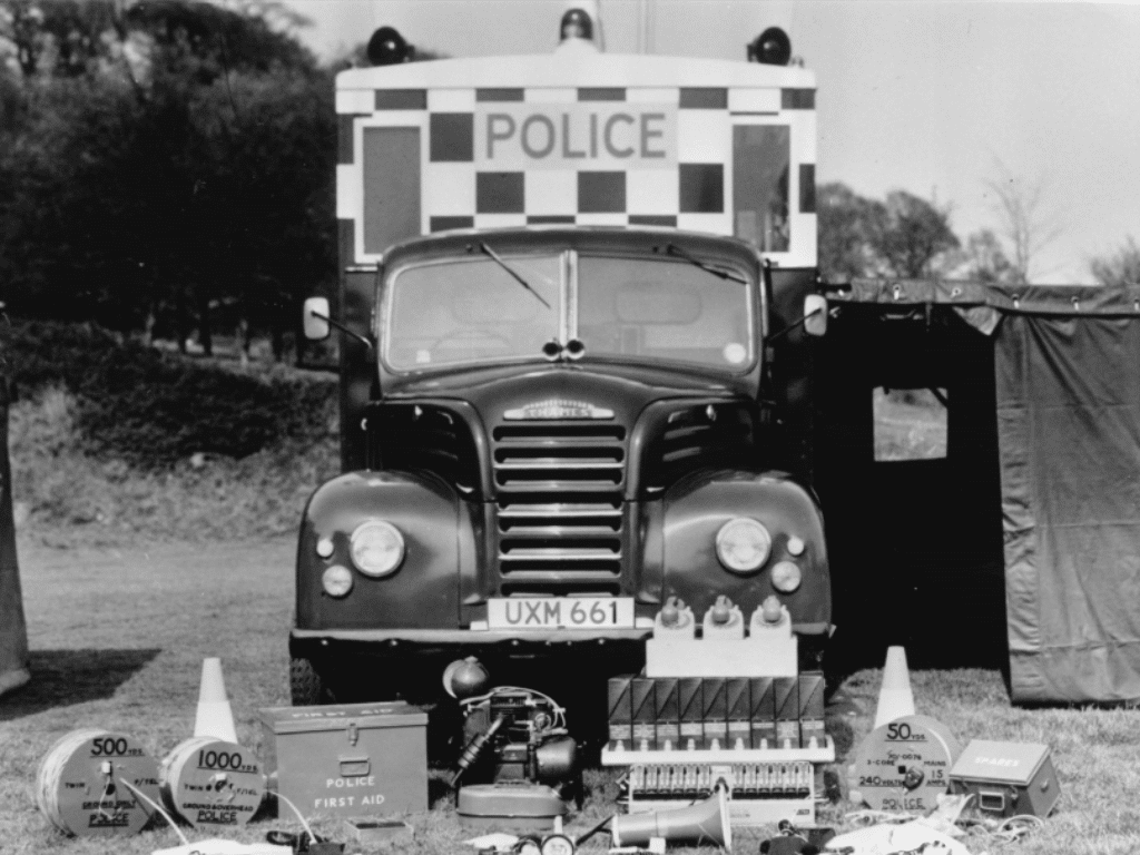 Black and white image of the first major incident police response vehicle.  The vehicle is parked in a field and this is the front view of it.  It has large police branding and a tent coming off the side. In front are police supplies on the grass.