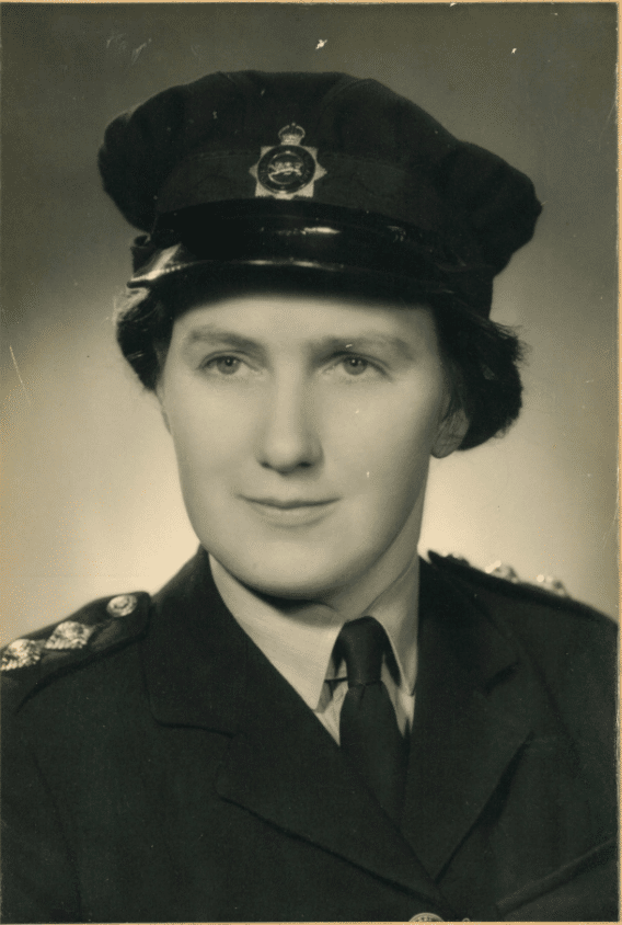 The image, which has sepia tones, shows a woman in a police hat, a jacket with epaulettes, and a shirt and tie. She is centralised in the frame, but looking just off camera with a slight smile on her face.