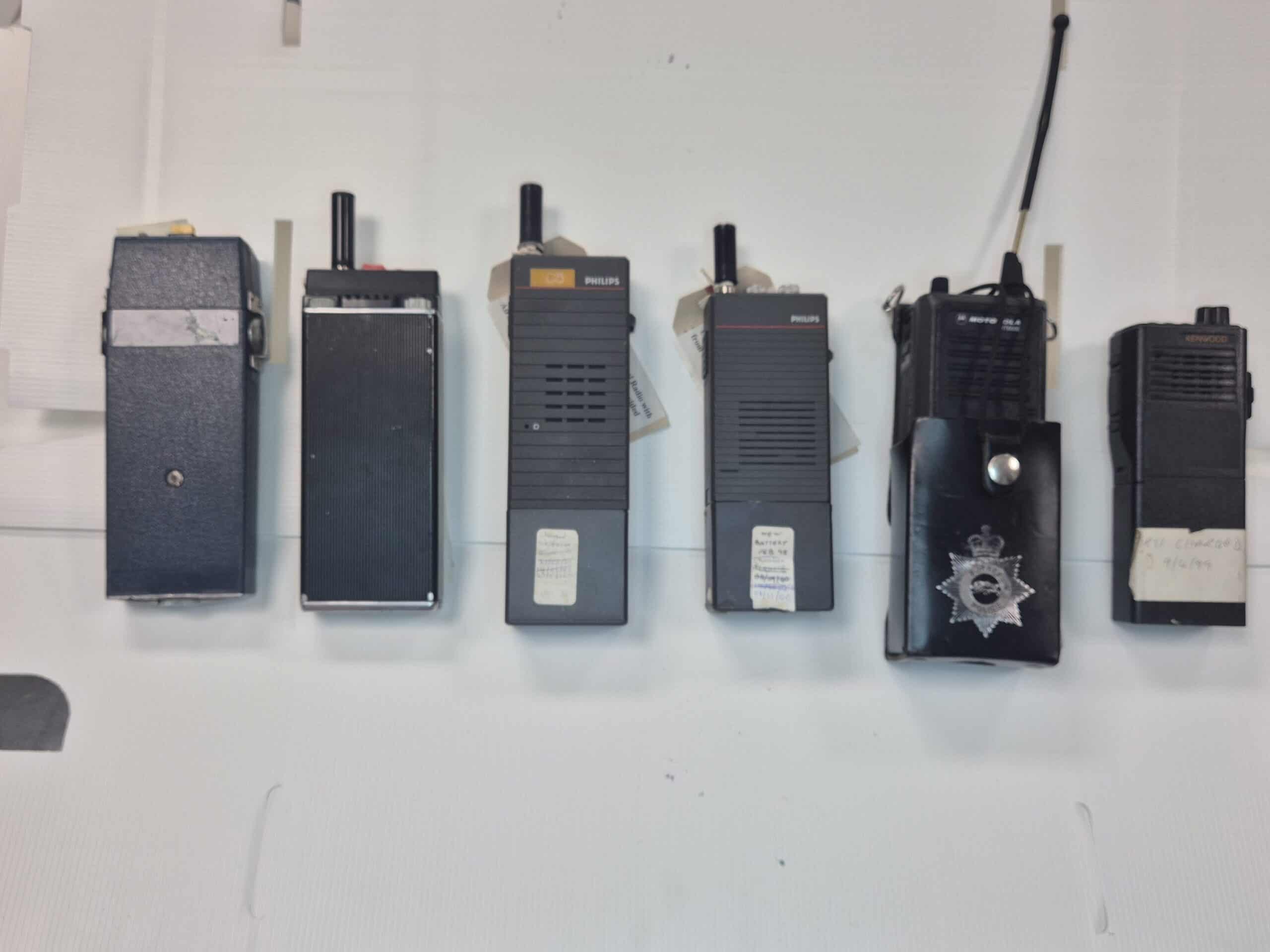 The image shows 6 black personal radios lined up on a table that were used by Surrey Police from 1960s to 2000s.  They are all of a similar size and the penultimate one on the far right is in a holister with the Surrey Police crest on it. 