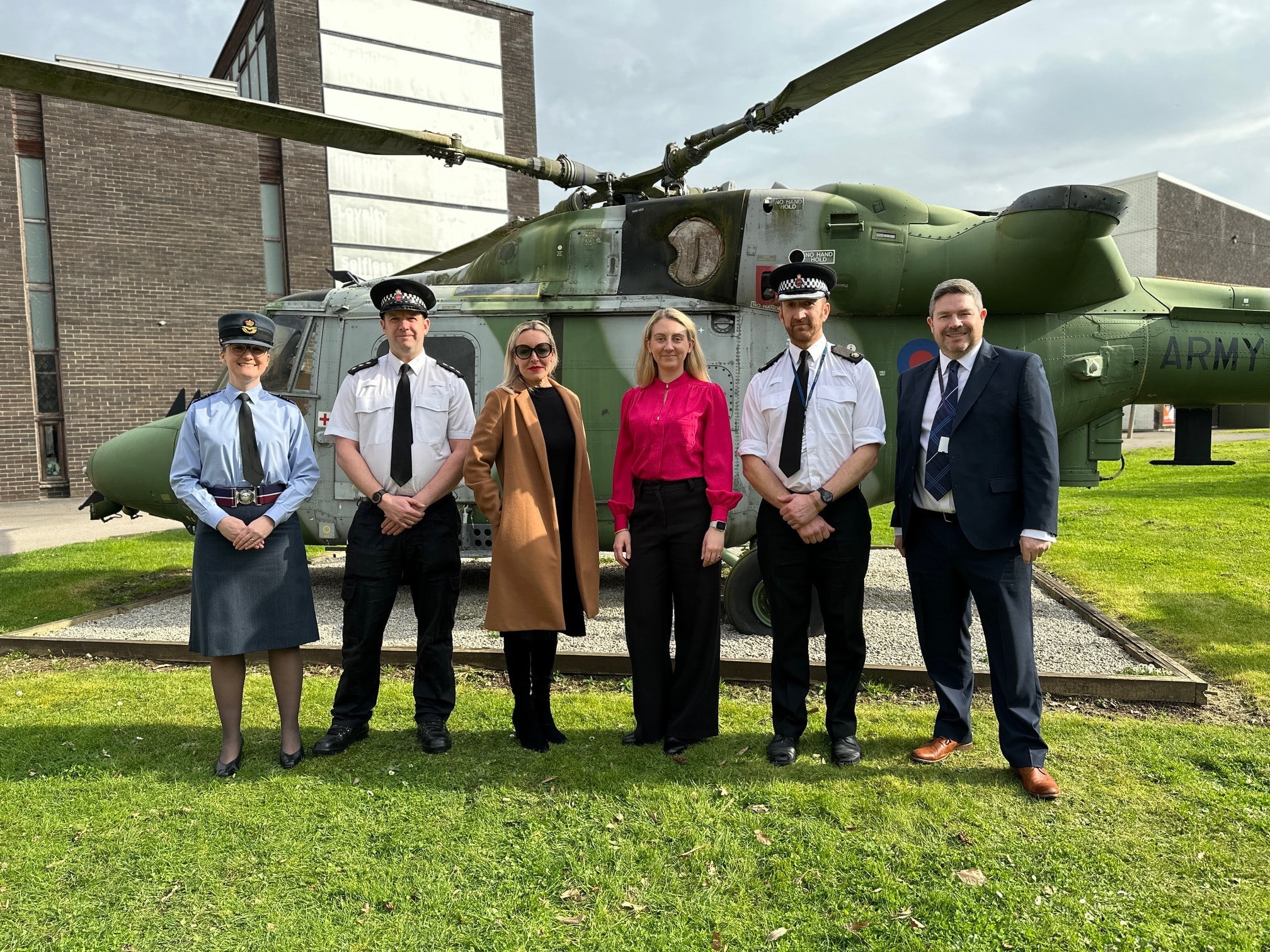 The image shows a group of six people standing in front of an Apache helicopter. Three of the six people pictured are in Surrey Police uniform. At the centre of the group, is Deputy Police and Crime Commissioner Ellie Vesey-Thompson, wearing pink, and Claire Lilly, who wears sunglasses and a tan coat. The image was taken at the Army Training Centre in Pirbright.
