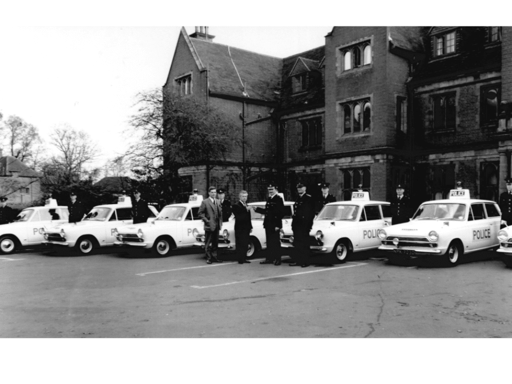 Black and white image of 7 white Ford Cortinas with the Police branding down the side of each vehicle and blue siren light on top.  The cars are lined up parked in front of the Old House at Mount Browne Headquarters in Guildford.  There is a police officer in uniform standing by the each of the cars and a couple of plain clothed gentlemen with two other officers in the centre of the image looking to camera.