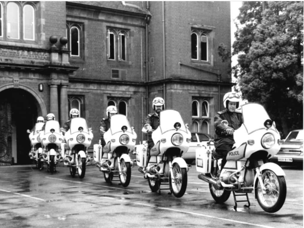 Black and white image of 6 white Police Honda motorcycles with Police branding on from 1970.  The motorcycles are in convoy one behind another outside Mount Browne Old House in Guildford.  Each motorcycle has a police officer in uniform riding it with white helmet and goggles.