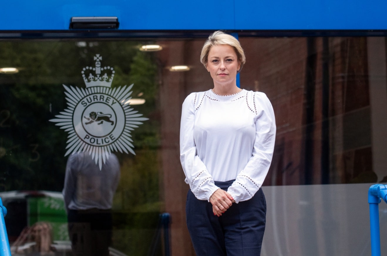 The image shows Police and Crime Commissioner Lisa Townsend, a blonde woman who wears a white long-sleeved top and black trousers, standing close to a glass door which has the Surrey Police logo on it. Her hands are clasped and she is looking directly into the camera with a serious expression.